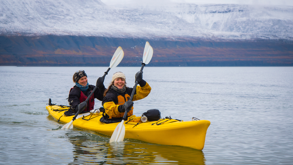 Two smiling people in a kayak in front of snowy mountains