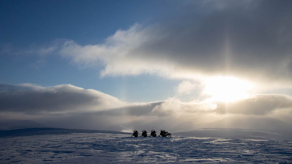 Four snowmobiles on the snow while the sun shines through the clouds.